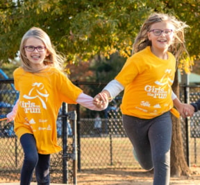 Two Girls on the Run participants in yellow shirts run while holding hands and smiling